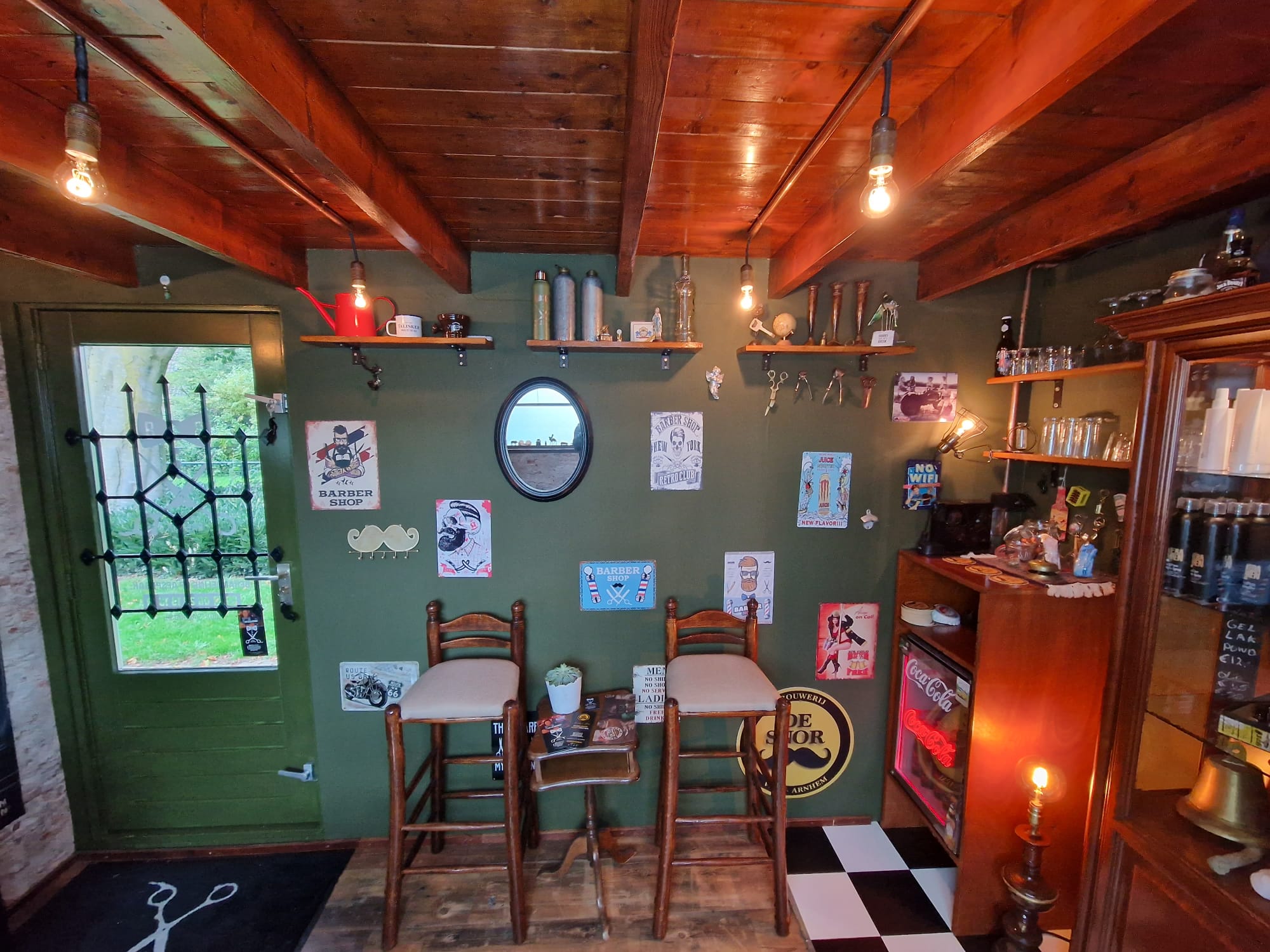 De salon van Bas Barber: groene wand, houten balken, twee barkrukken en een zwart-witte tegelvloer